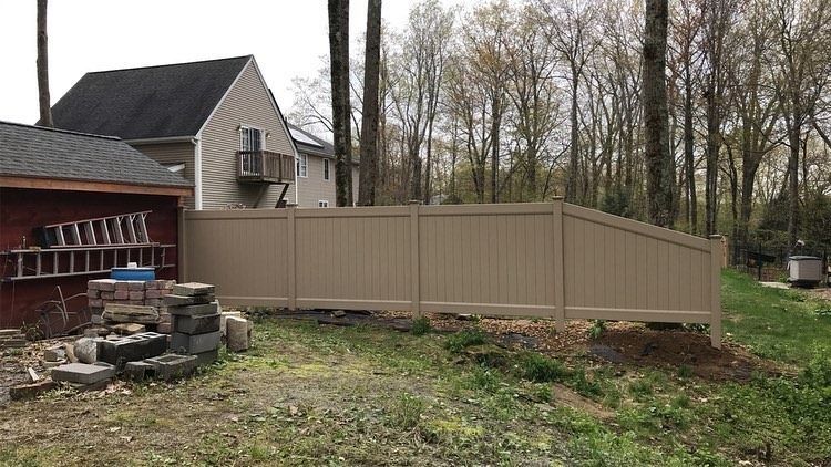 A tan vinyl privacy fence stands in a backyard, partially obscuring a house and a wooded area in the background.