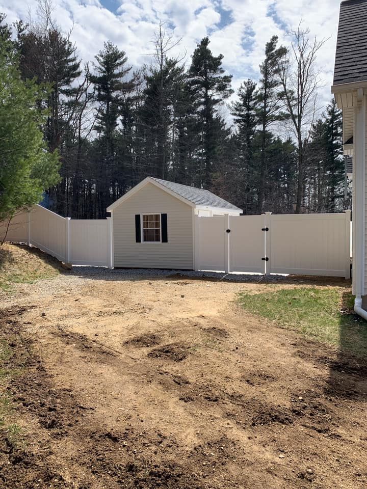 A small beige shed with a black-shuttered window sits behind a white vinyl fence in a dirt-covered backyard.