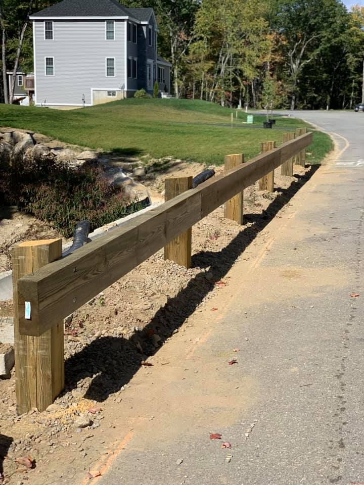 A wooden guardrail runs along the edge of a paved road next to a grass hill and a gray house in the background.