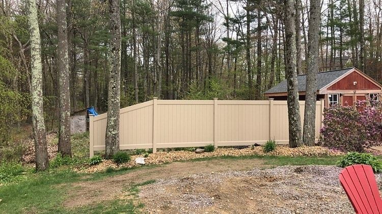 A beige vinyl fence spans a backyard, with tall trees in the background and a small red shed visible on the right.