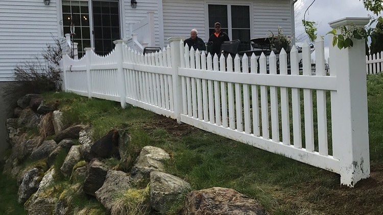 A white vinyl picket fence runs along the top of a stone retaining wall in front of a white house.