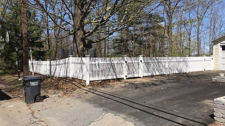 A white picket fence encloses a backyard bordering a wooded area, with a trash bin in the foreground on an asphalt drive.
