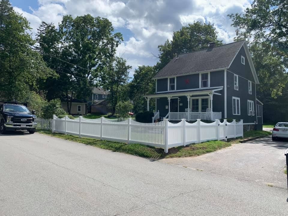 A dark gray two-story house with a white porch and a surrounding white picket fence under a partly cloudy sky.