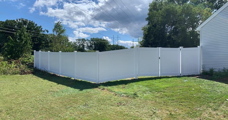 A white vinyl privacy fence encloses a green grassy backyard next to the side of a white house under a blue sky.