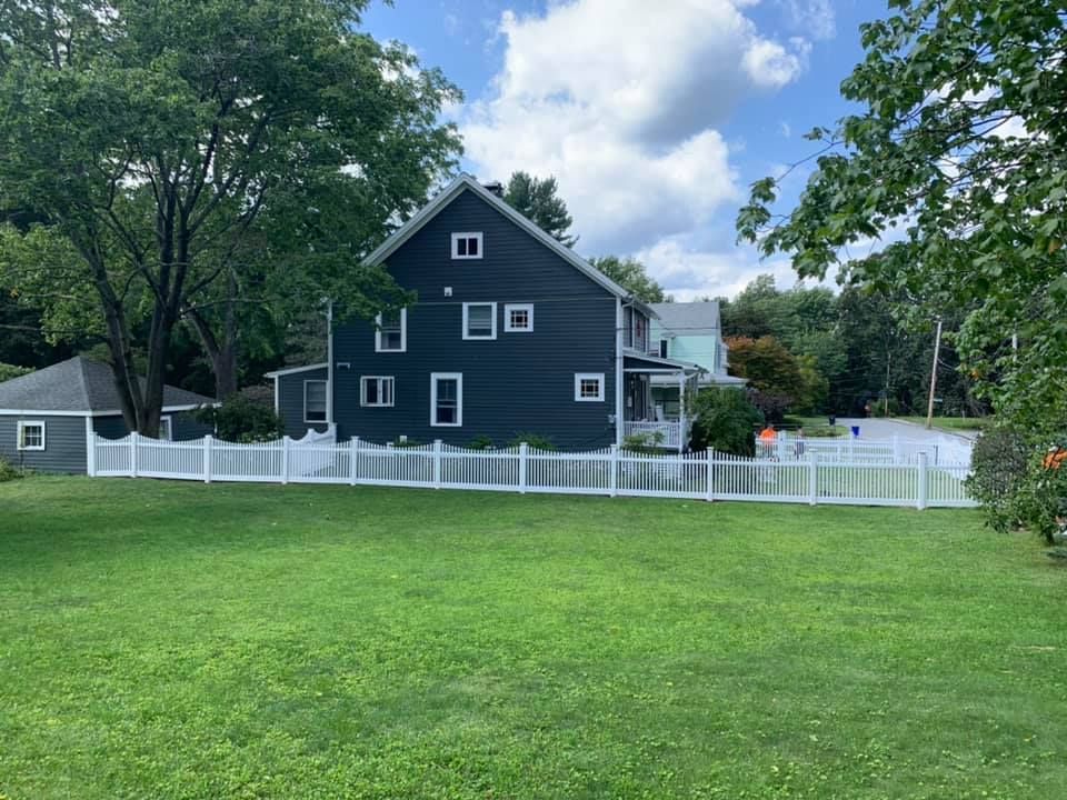 A dark blue two-story house with a white picket fence, surrounded by green grass and trees under a blue sky.