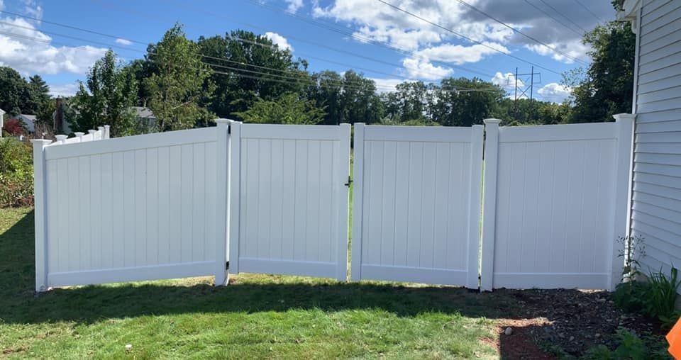 A white vinyl privacy fence with a double gate stands on a grassy lawn under a blue, cloudy sky.