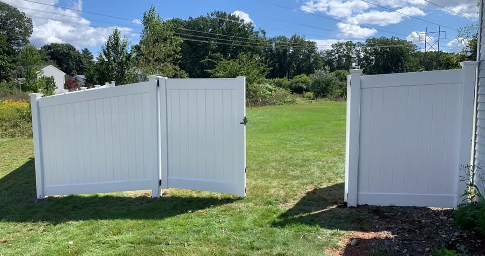 A white vinyl fence with a double gate standing open in a grassy backyard under a bright blue sky.
