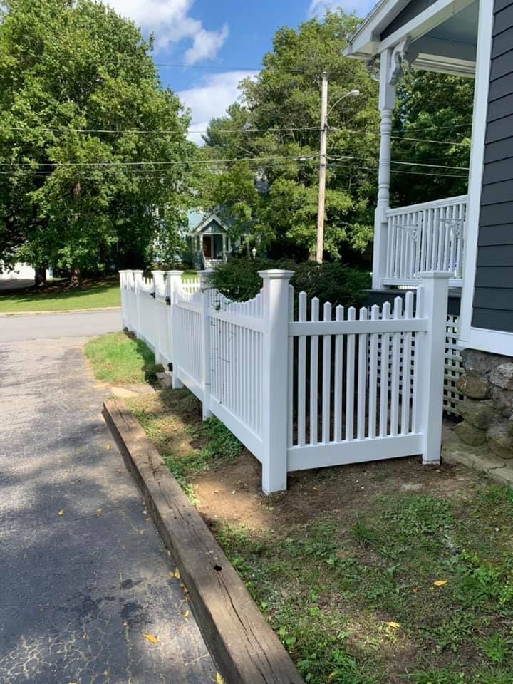 A white vinyl picket fence extends from the side of a house toward a driveway, with trees and a distant home in the back.