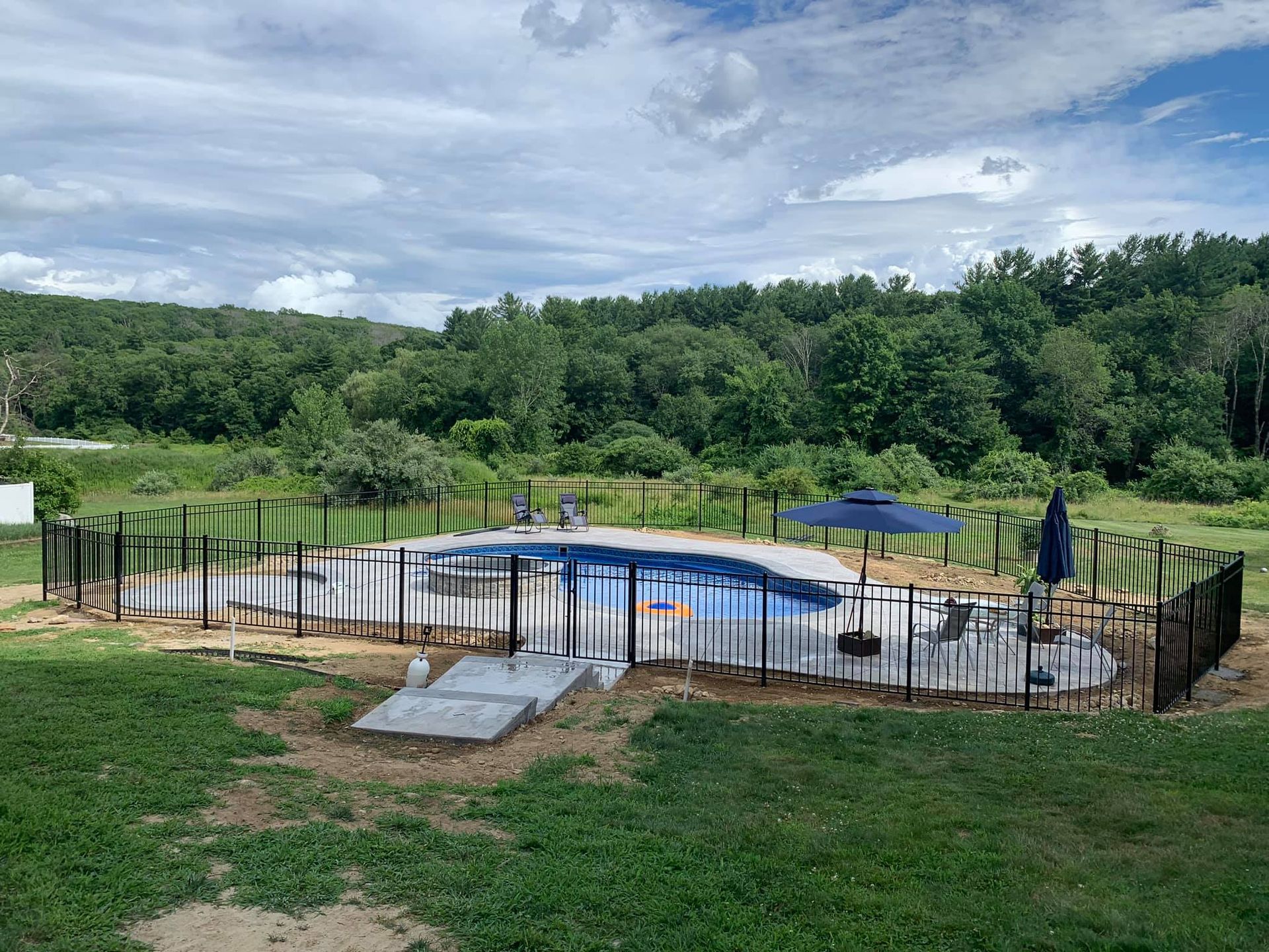 An outdoor swimming pool surrounded by a black fence and a grassy yard, with a blue umbrella on a patio.