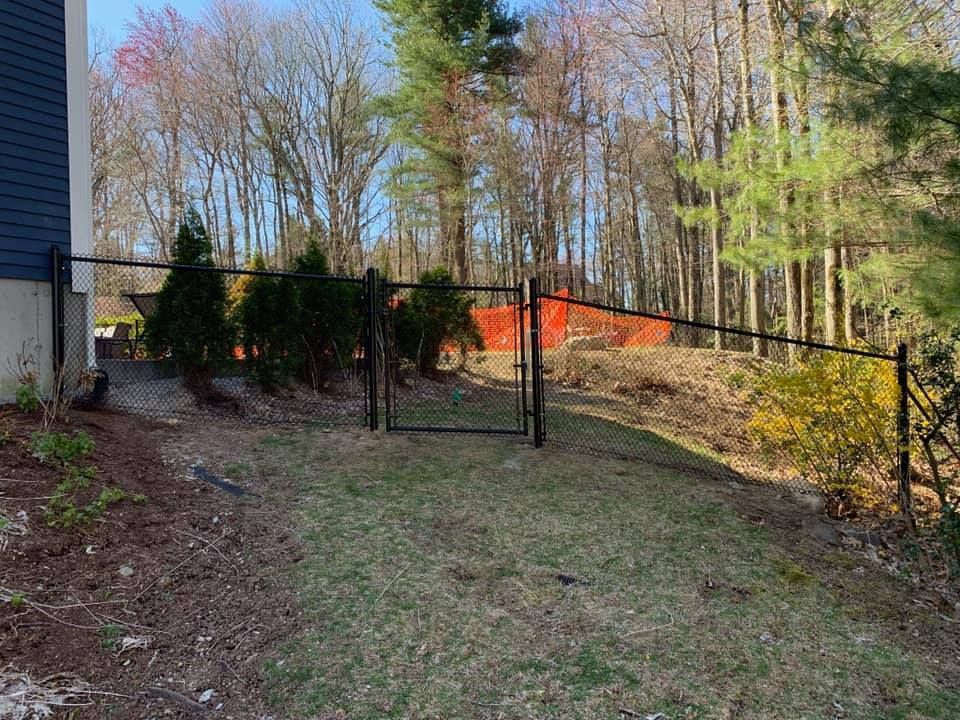 A black metal chain-link fence with a gate stands in a backyard, leading toward a forest with orange construction mesh.