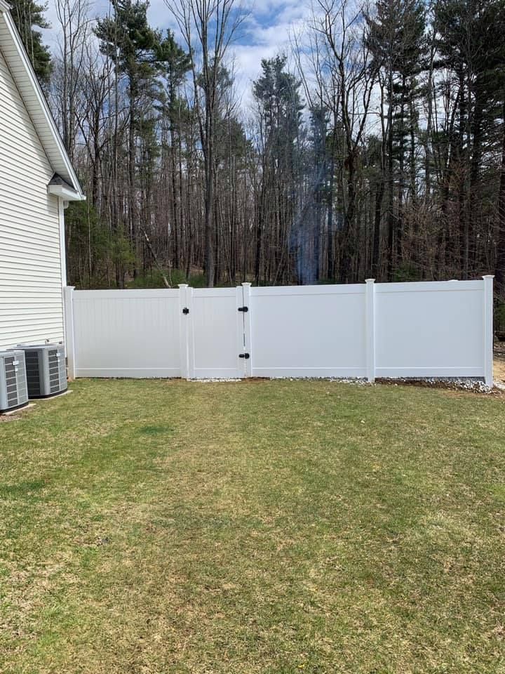 A white vinyl privacy fence with a closed gate sits in a residential backyard, bordering a wooded area.