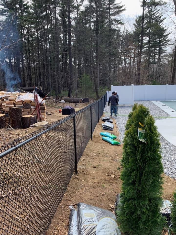 A person walks along a black chain-link fence bordering a wooded yard with cut logs and a partial view of a white fence.