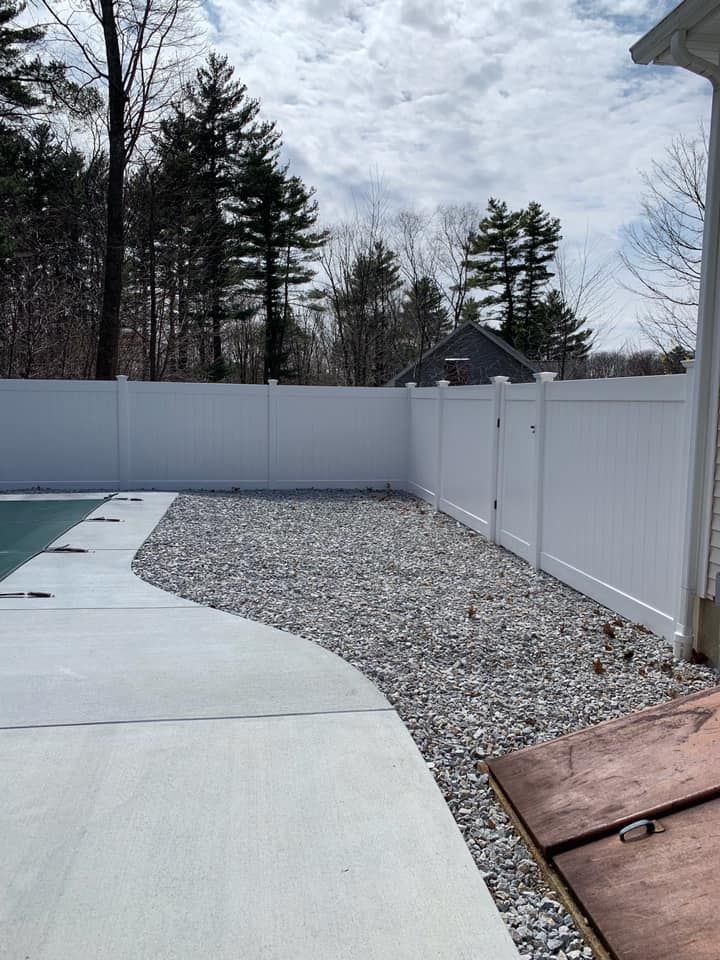 A white vinyl fence borders a backyard with a concrete pool deck and a gravel area, set against a backdrop of trees.