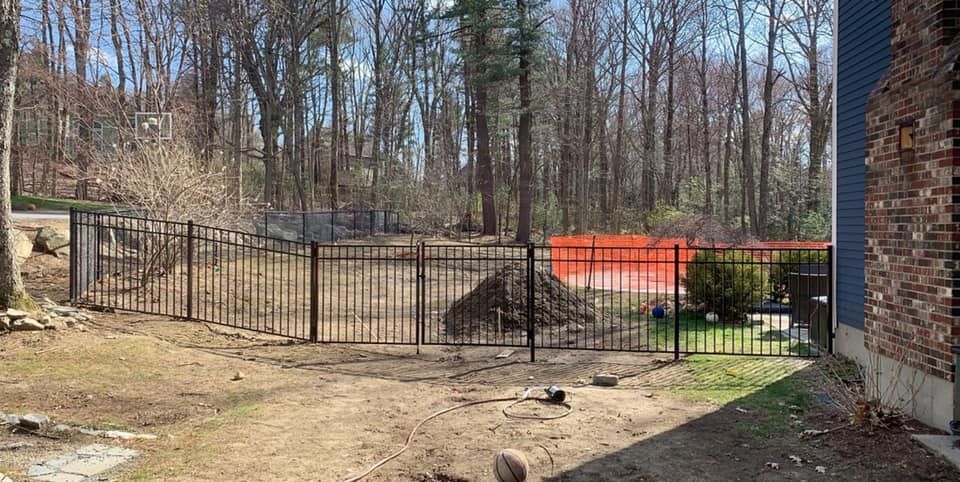 A backyard view with a metal fence, a pile of dirt, and an orange safety net in front of a wooded area.