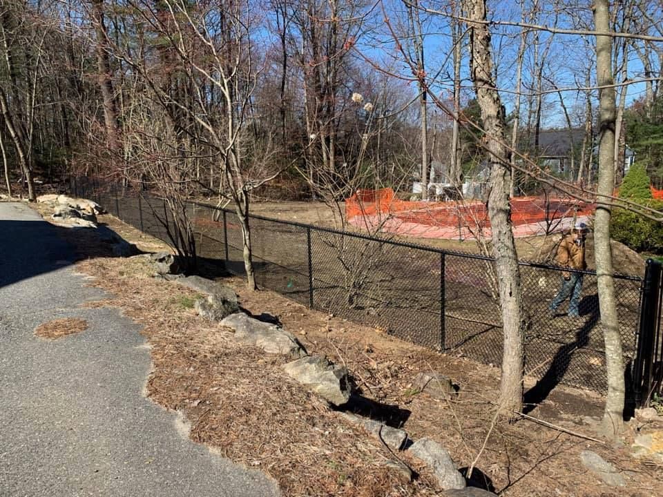 A chain-link fence lines a dirt lot with orange construction netting in the background, near a paved path and bare trees.