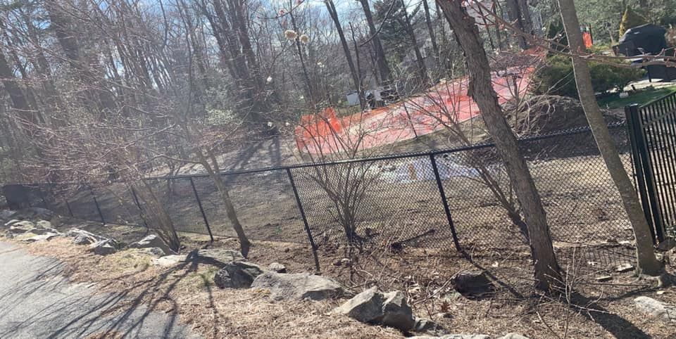 A black chain-link fence divides a foreground of dry leaves and rocks from a wooded area with orange construction fencing.