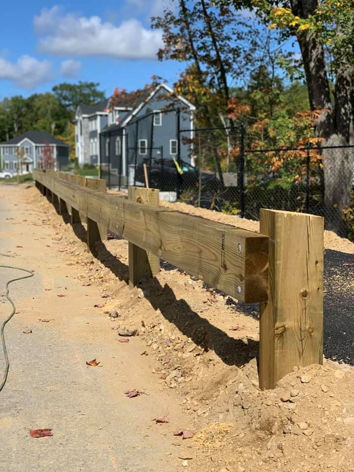 A wooden guardrail made of thick beams runs along a dirt roadside next to a grassy area, trees, and houses in the distance.