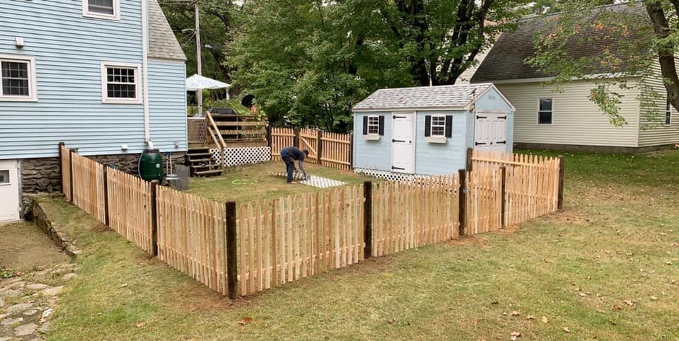 A person works in a fenced backyard containing a blue shed next to a light blue house on a grassy lot.