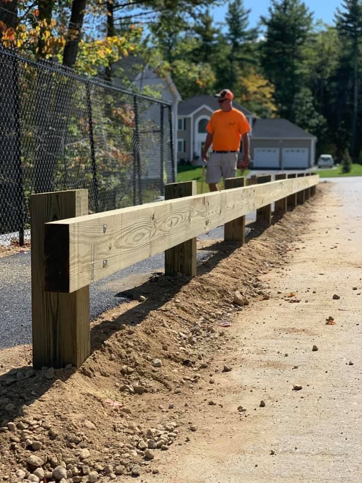 A person in an orange shirt stands next to a newly installed wooden post-and-rail fence along a dirt path.