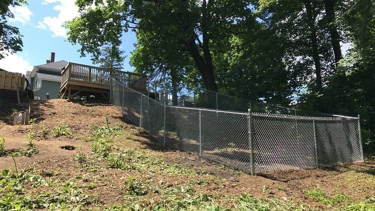 A chain-link fence encloses a sloped, cleared backyard area next to a wooden deck on a sunny day.