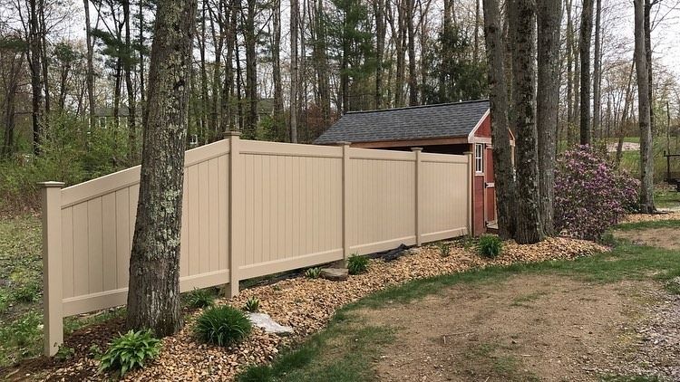 A tan privacy fence stands in a wooded backyard next to a small red shed with a dark roof and a bed of landscaping stones.