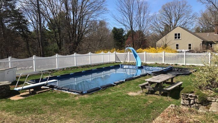 A backyard swimming pool with a blue cover, a slide, and a diving board, enclosed by a white picket fence.