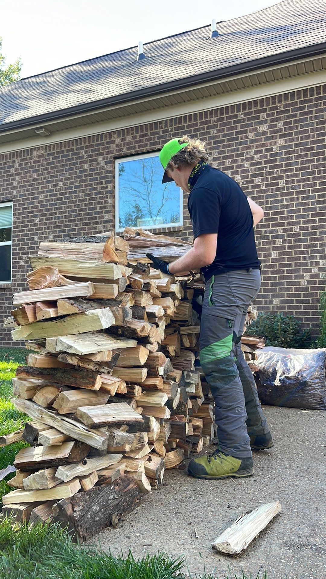 Person stacking cut firewood beside a brick house on a driveway