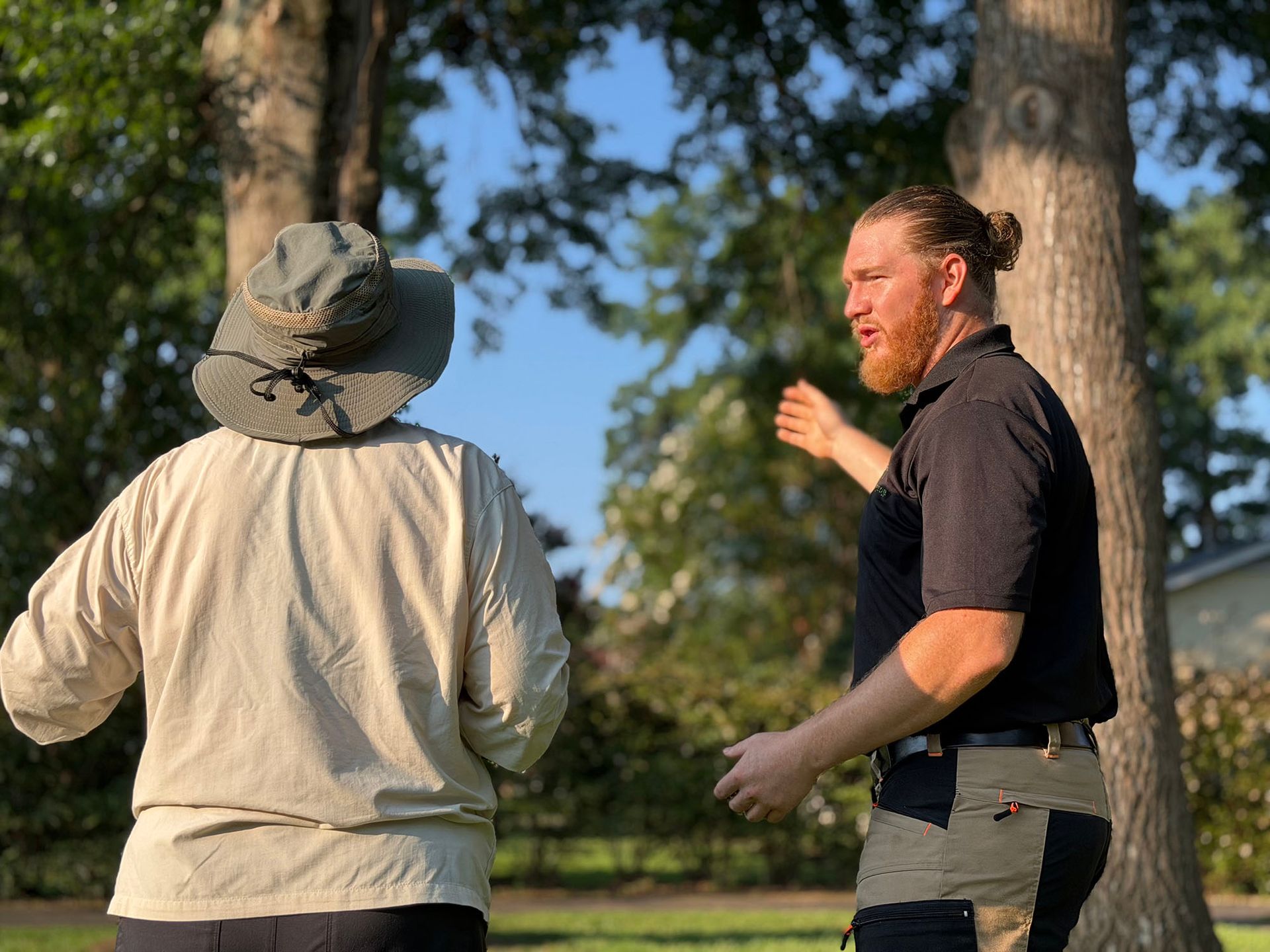 Two people outdoors among trees, one pointing while facing the other in a hat.