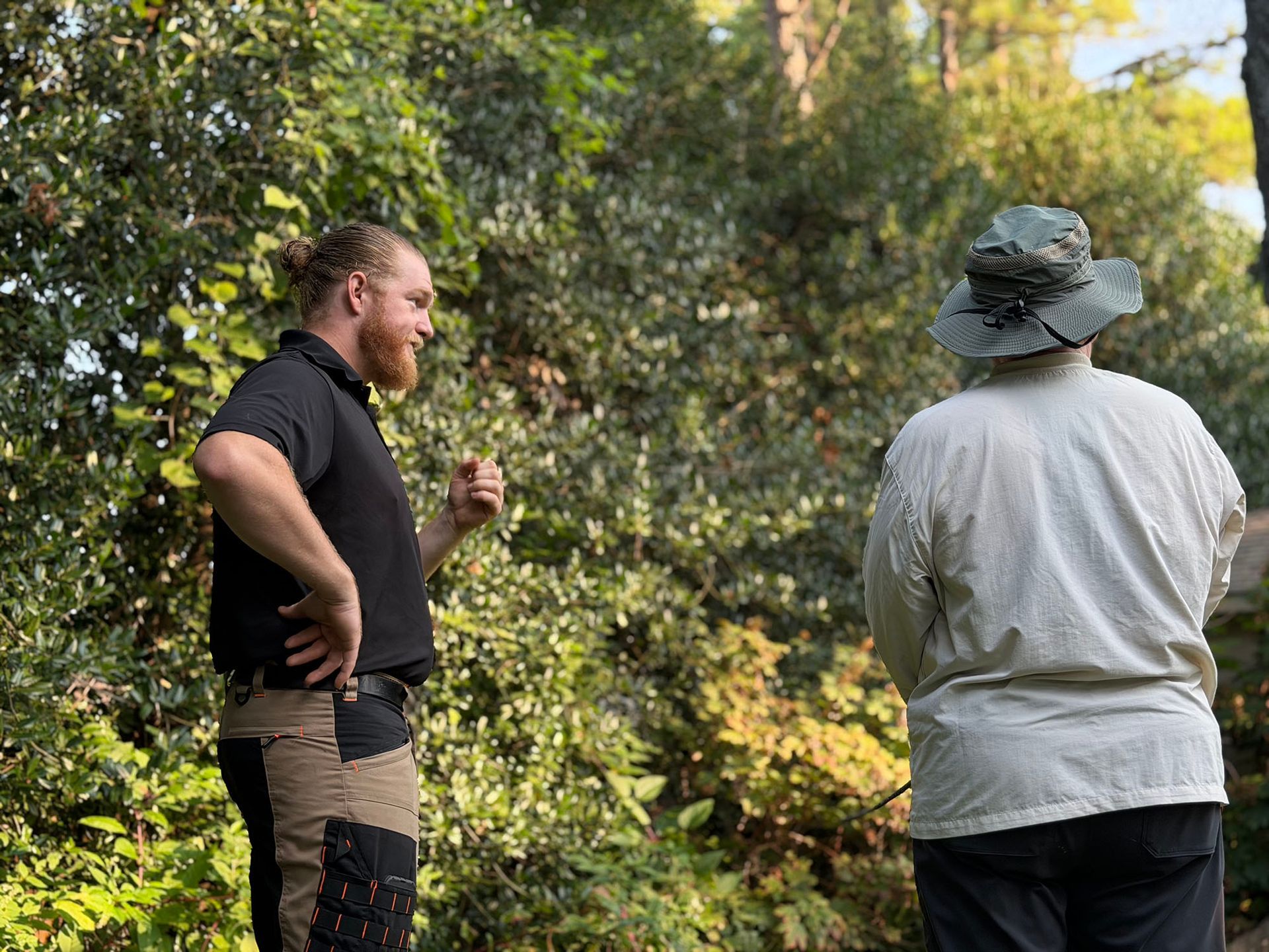 Two people standing and talking outdoors among trees, one in black and one in a light jacket with a hat.