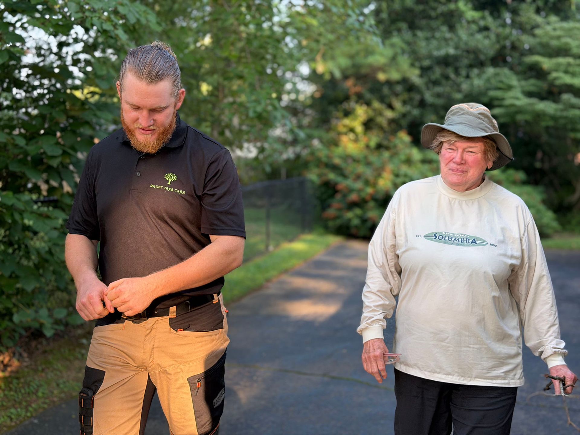 Two people walking on a tree-lined path, one in a black shirt and tan pants, the other in a light long-sleeve shirt.