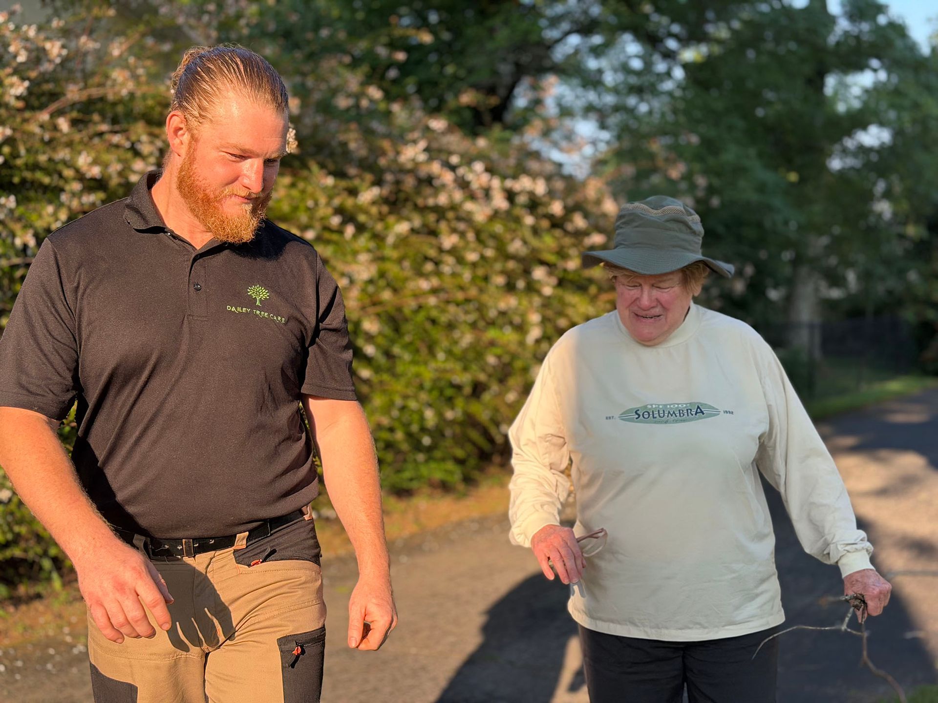 Two people walking outdoors on a sunny path, one carrying a leash, with trees and flowers in the background