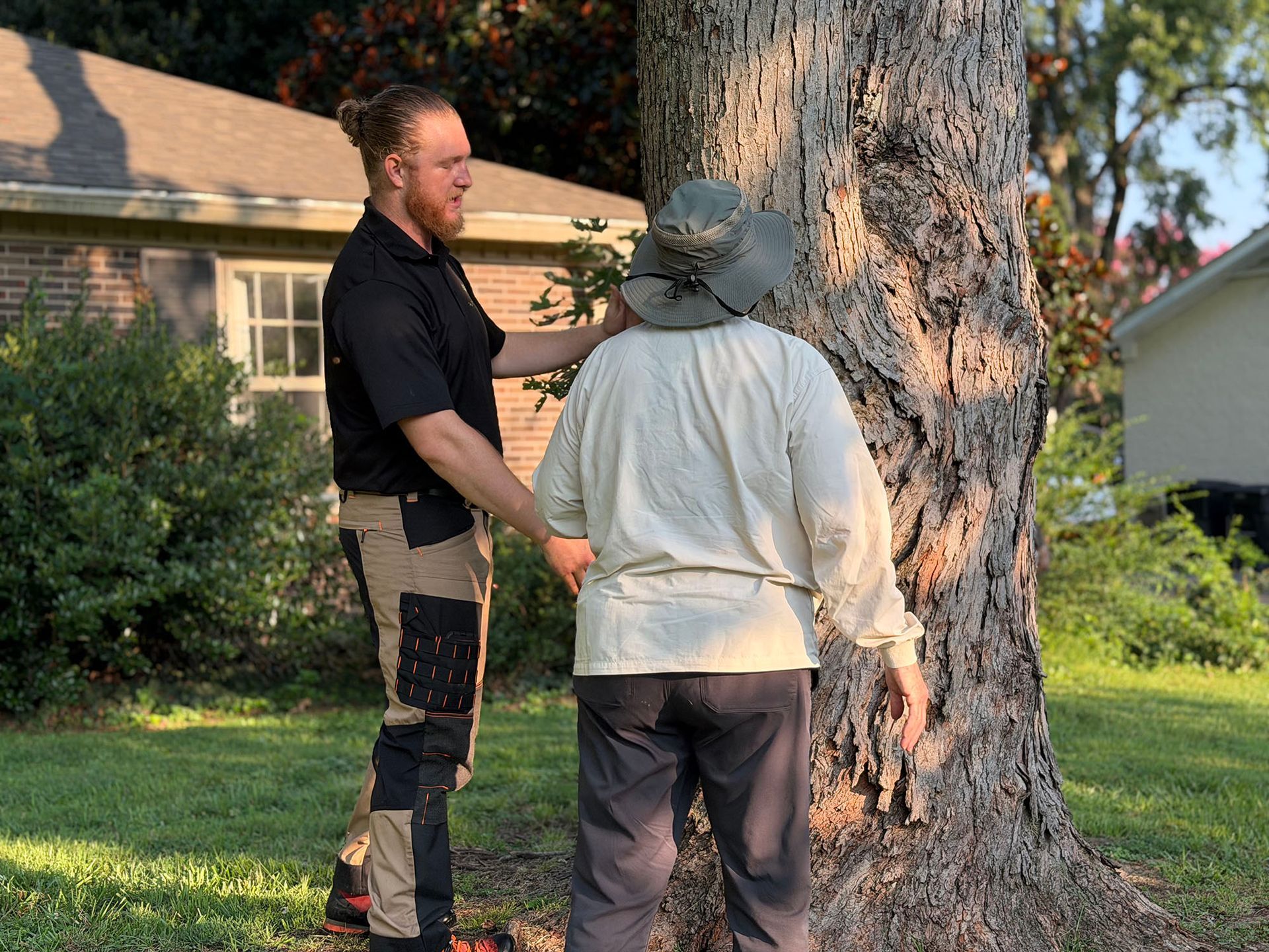 Two people stand by a large tree in a suburban yard, one facing away and one in black clothing.