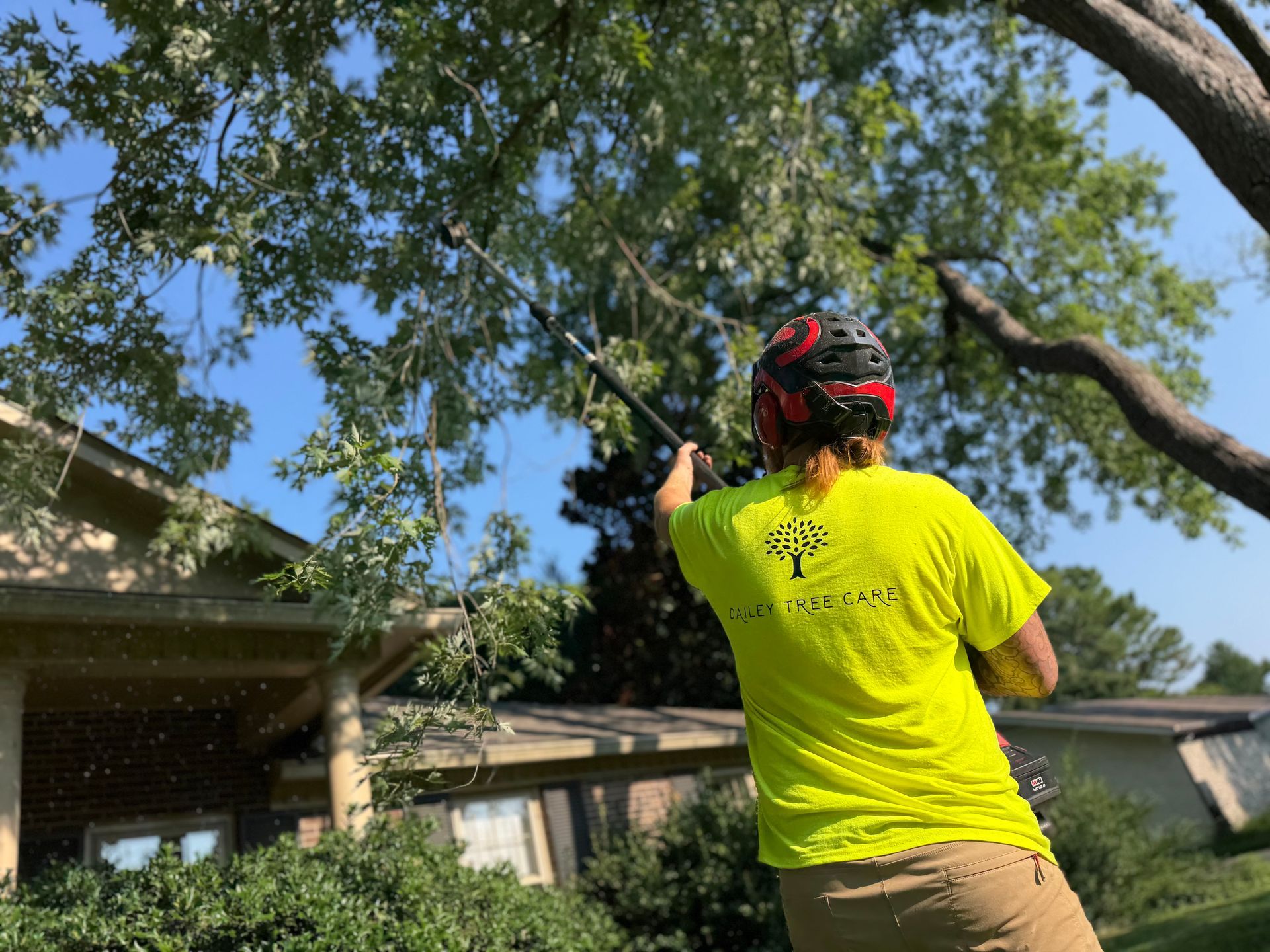 Person in neon yellow shirt and red helmet aiming a bow in a backyard under a tree.