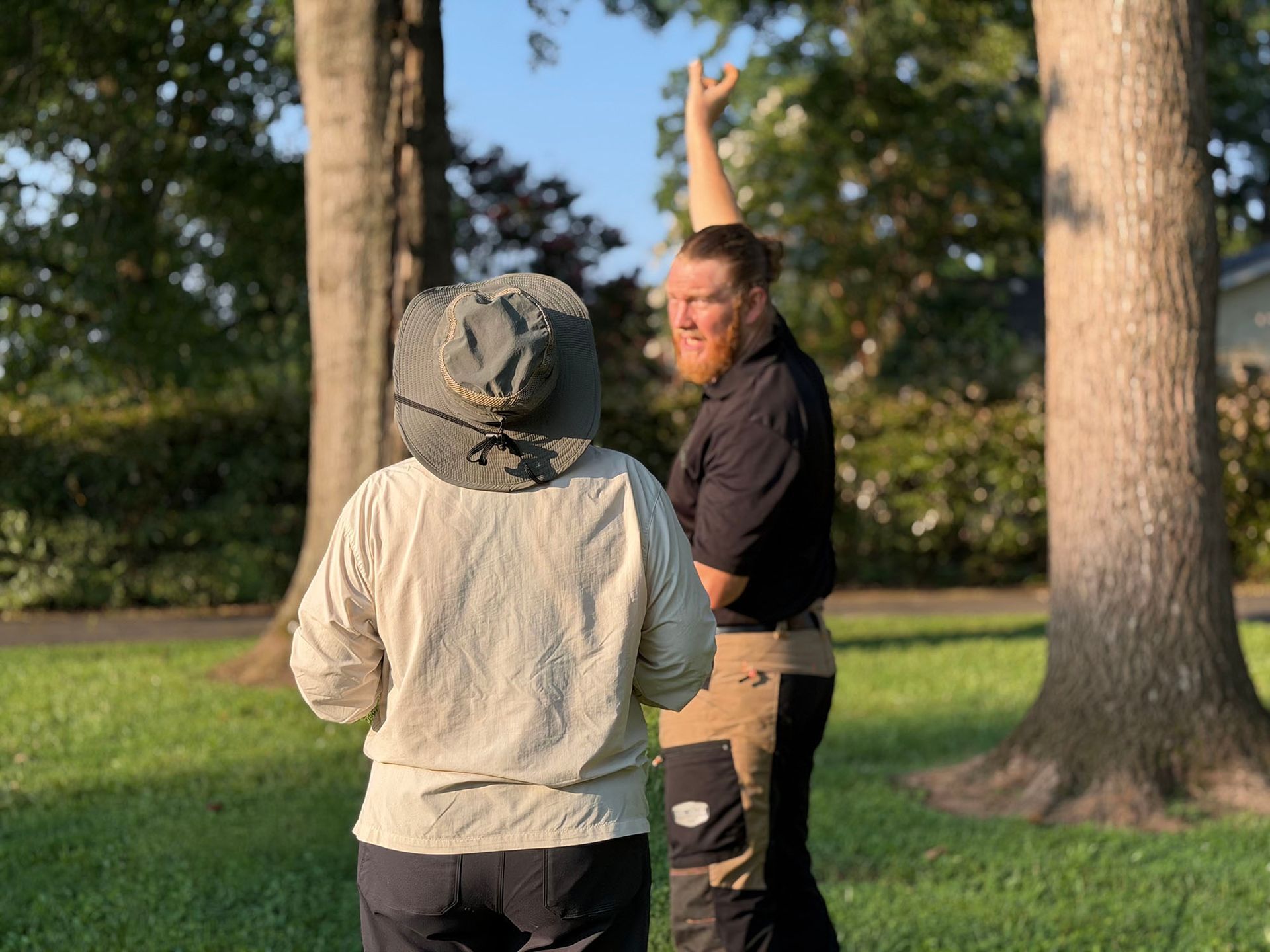 Two people outdoors in a grassy park, one raising an arm while facing the other near tall trees.