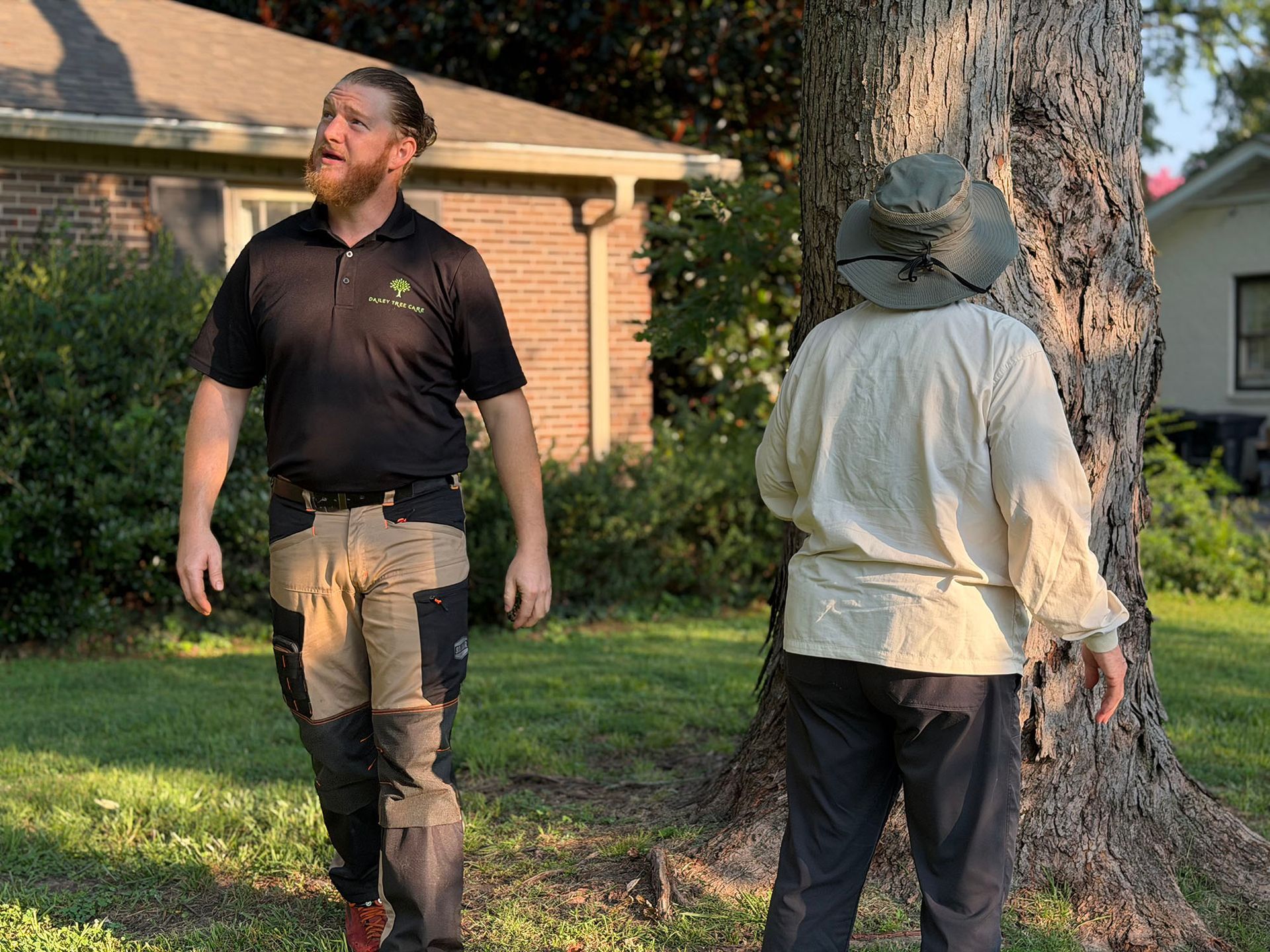 Two people stand outdoors by a tree in a yard, one facing left and one facing away.