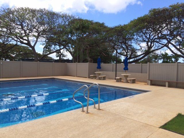 Swimming pool surrounded by a beige fence, picnic tables, and trees against a partly cloudy sky.