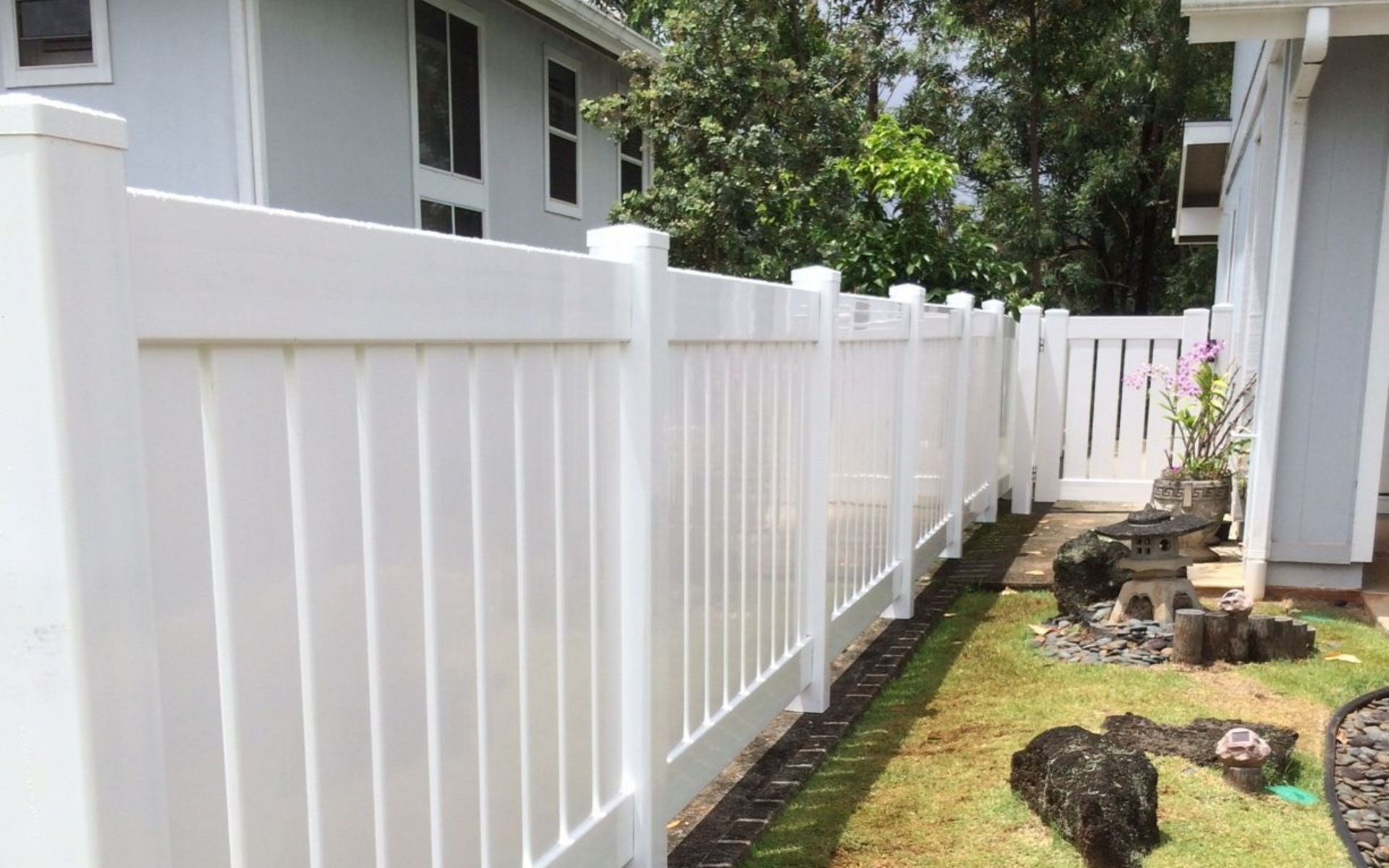 White vinyl fence in a yard with grass, rocks, and houses in the background.