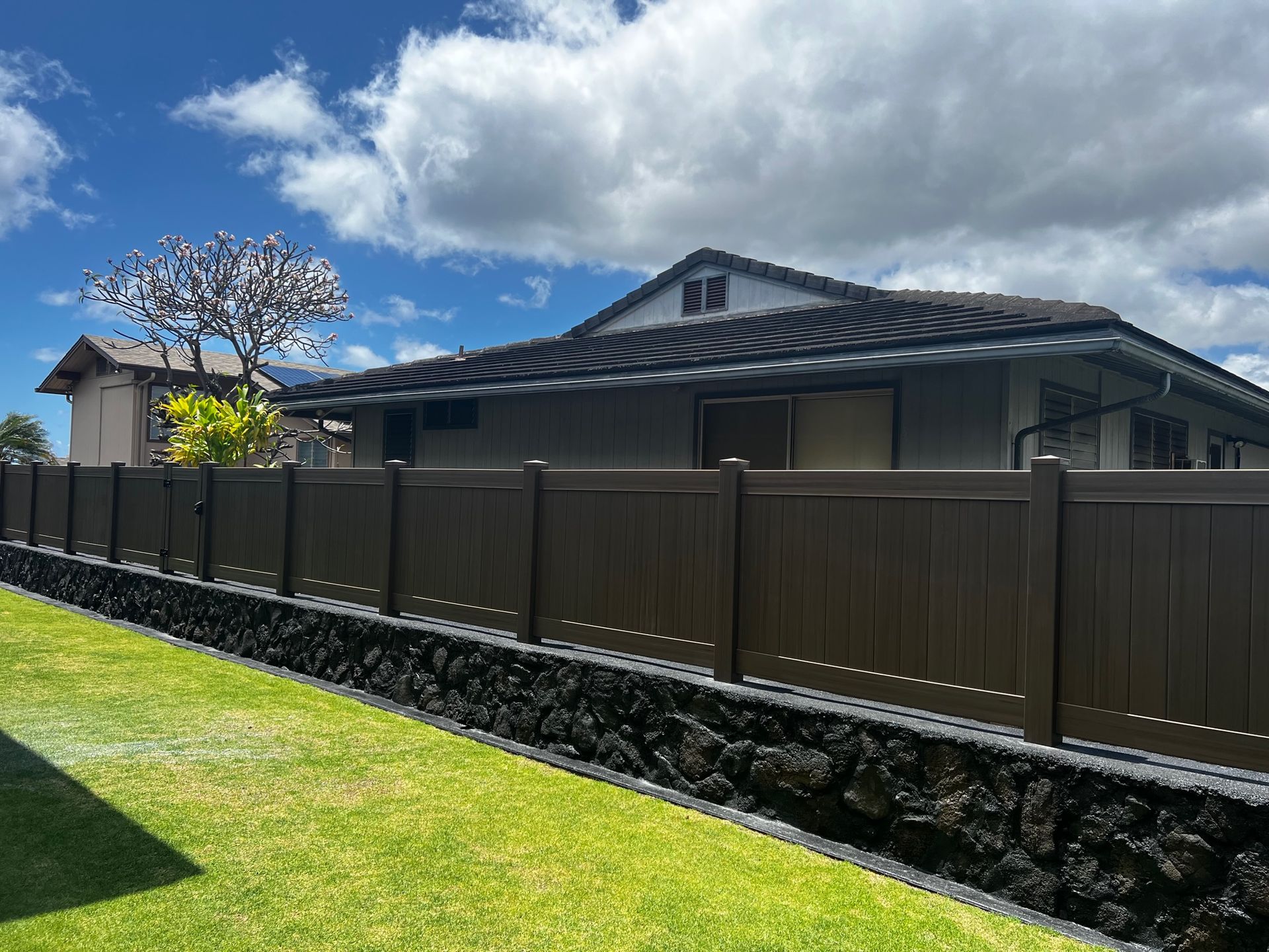 Brown fence on a black rock wall along a green lawn with a house and blue sky in the background.