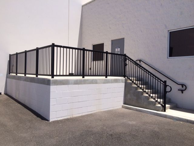 Black metal railing surrounds a raised platform with stairs and a handrail against a white building.