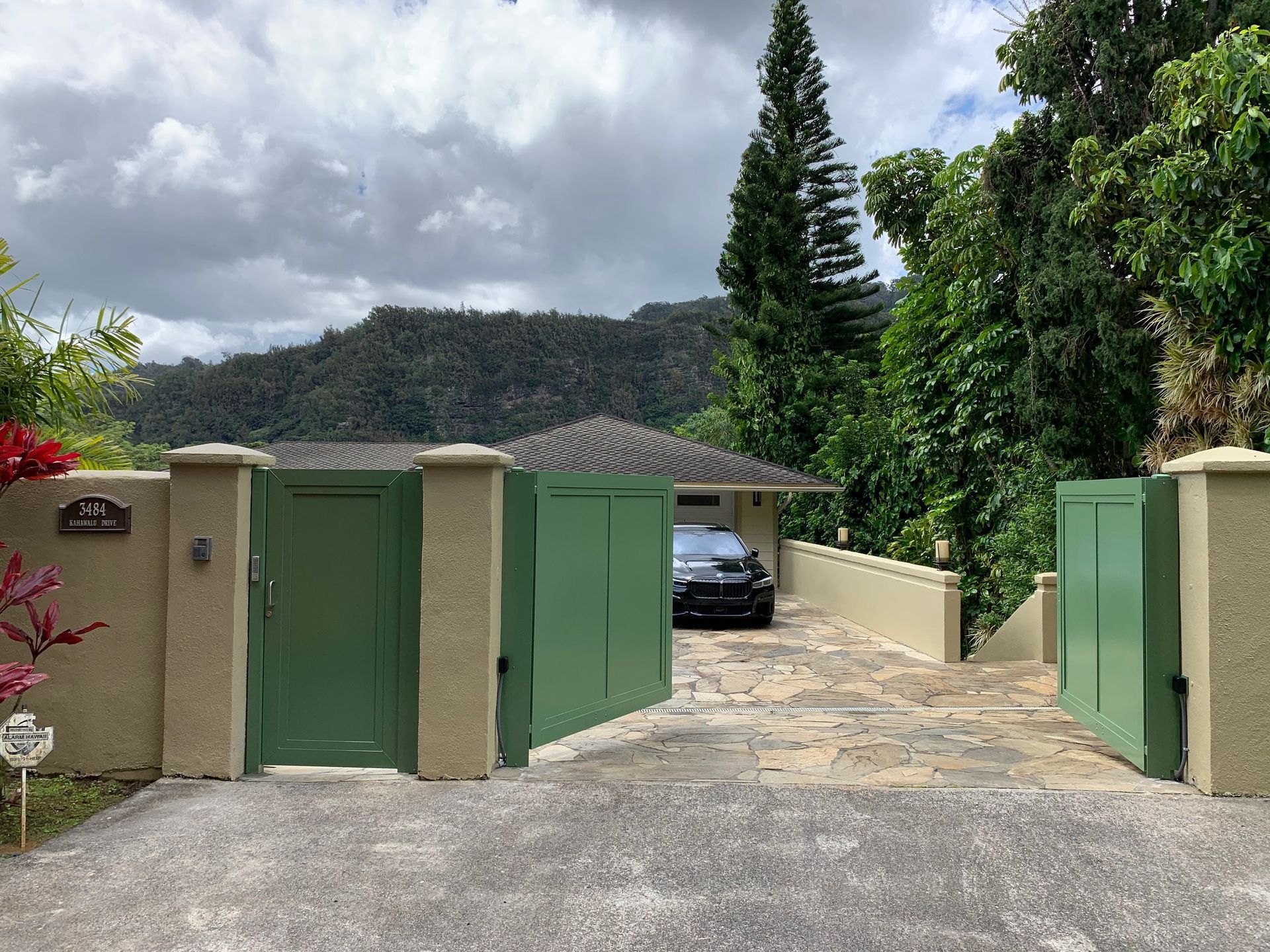House behind green gates; driveway, trees and mountains in the background. Cloudy sky.