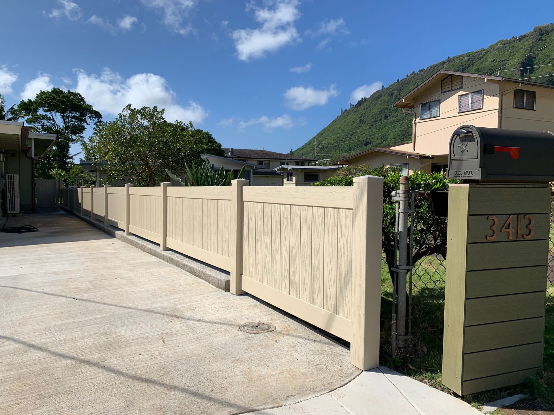 Tan wooden fence along a paved driveway with a house and mailbox in front of a green mountain under a blue sky.
