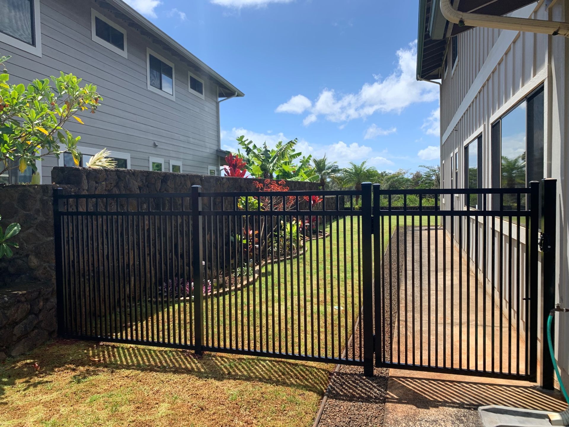 Black metal fence and gate in front of a yard, with two houses in the background on a sunny day.