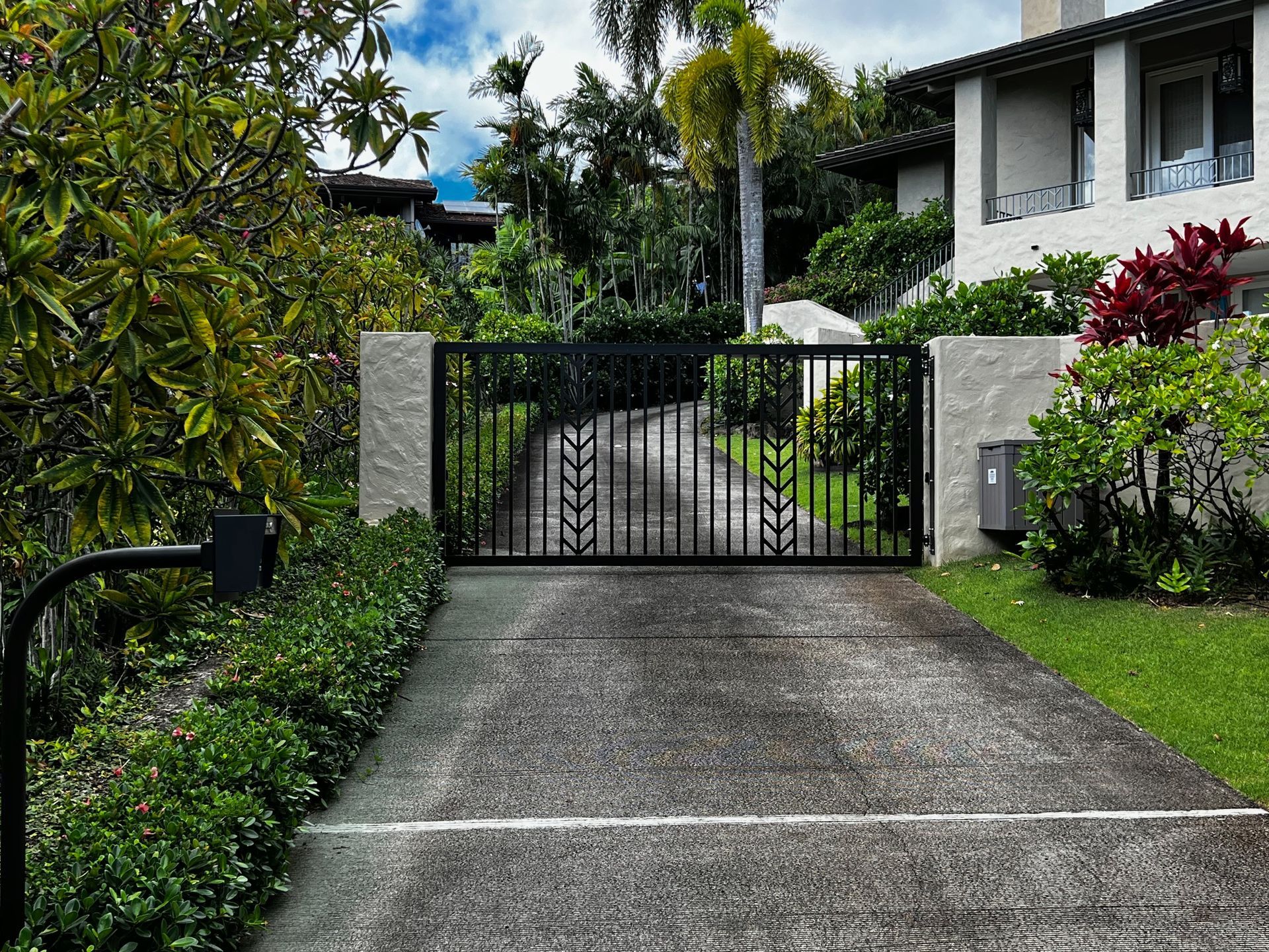 Black wrought-iron gate in front of a driveway leading to a light-colored house, framed by stone pillars and greenery.