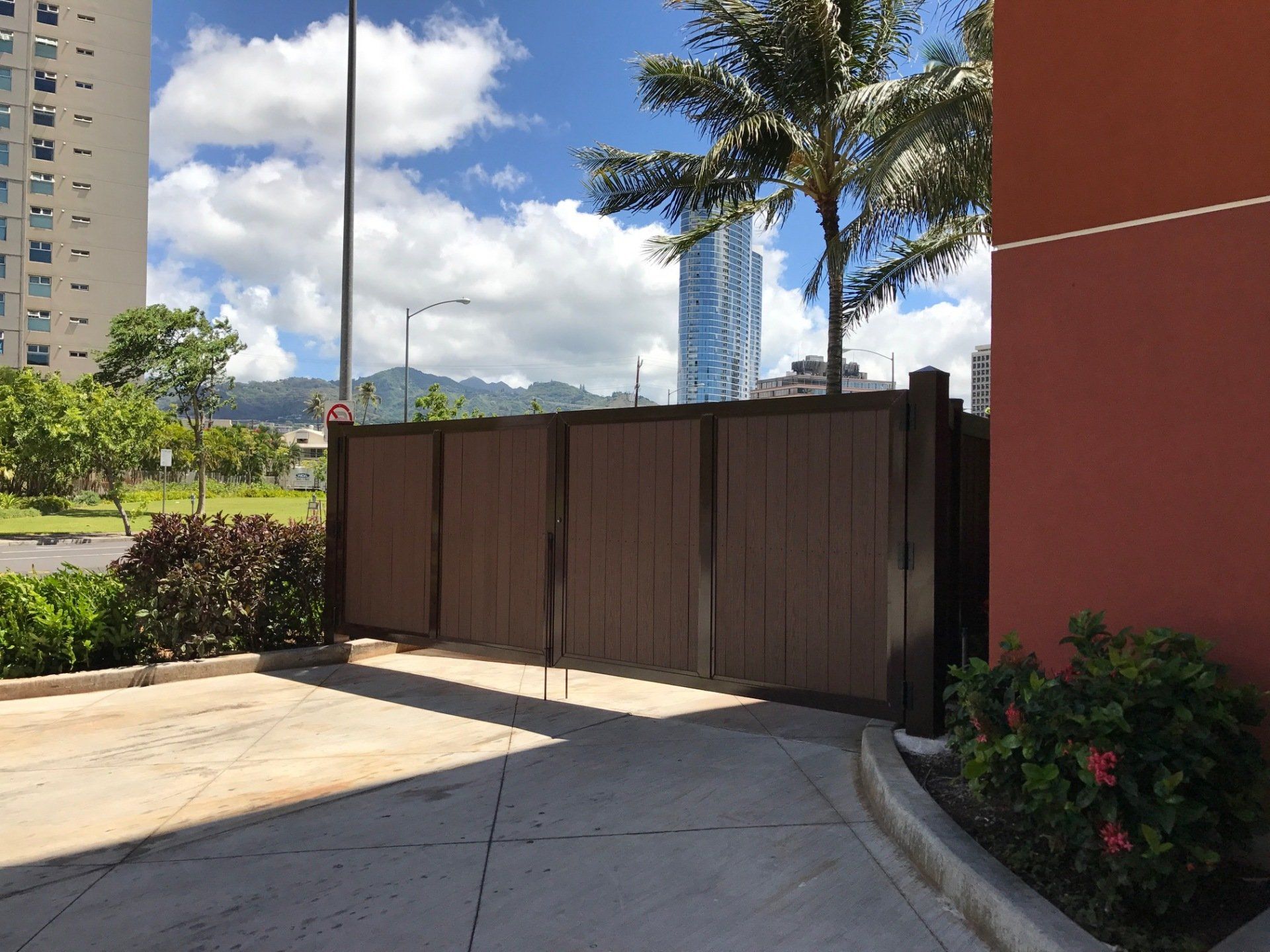 Brown gate blocks a driveway entrance. Tall buildings, blue sky, and a palm tree are visible in the background.