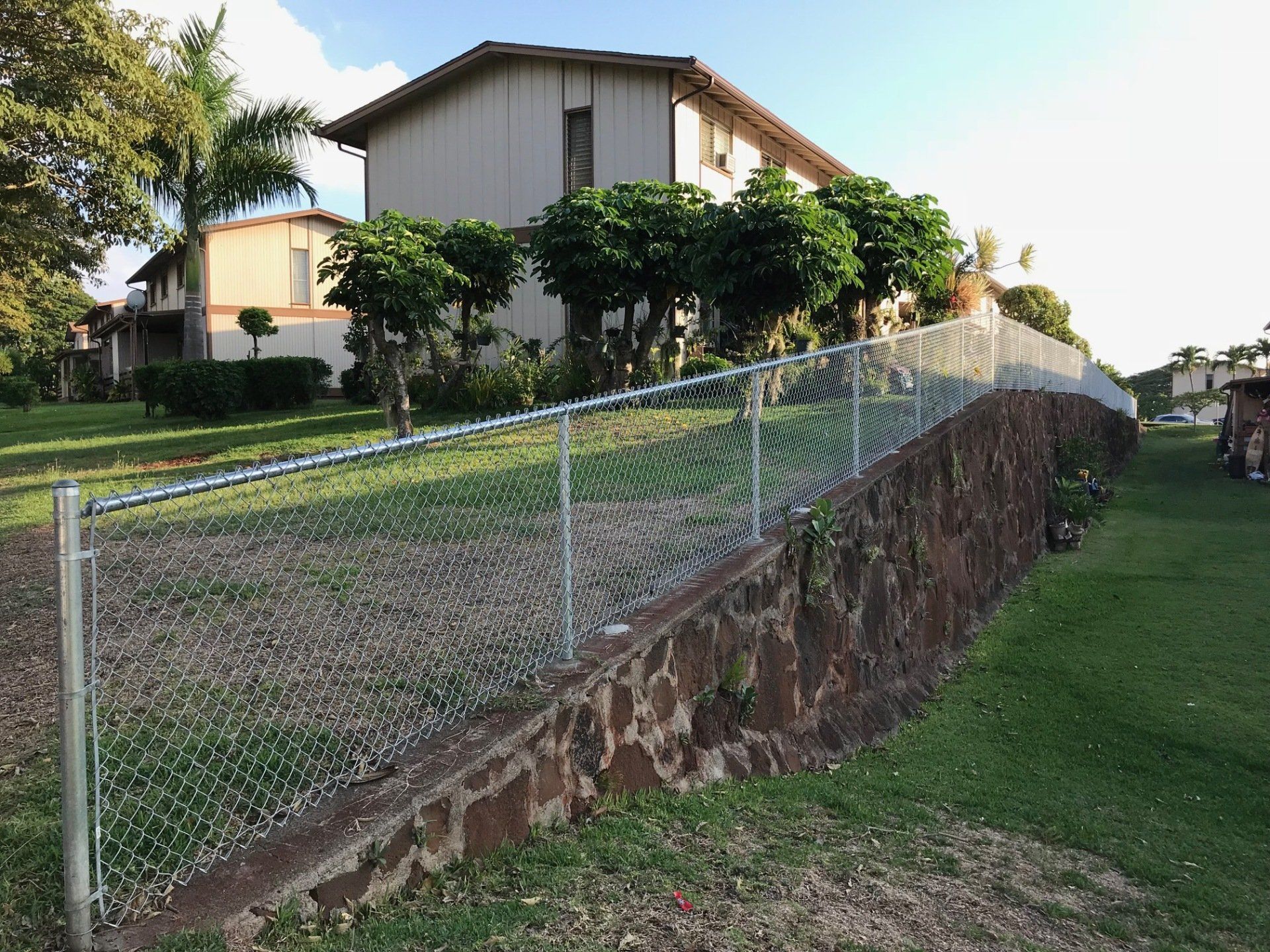 Chain-link fence atop a stone retaining wall borders a grassy area. Two-story building in the background.