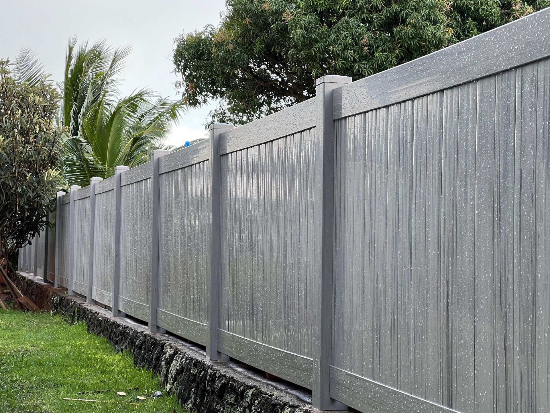 Gray speckled privacy fence along a stone border with green grass and trees in the background.