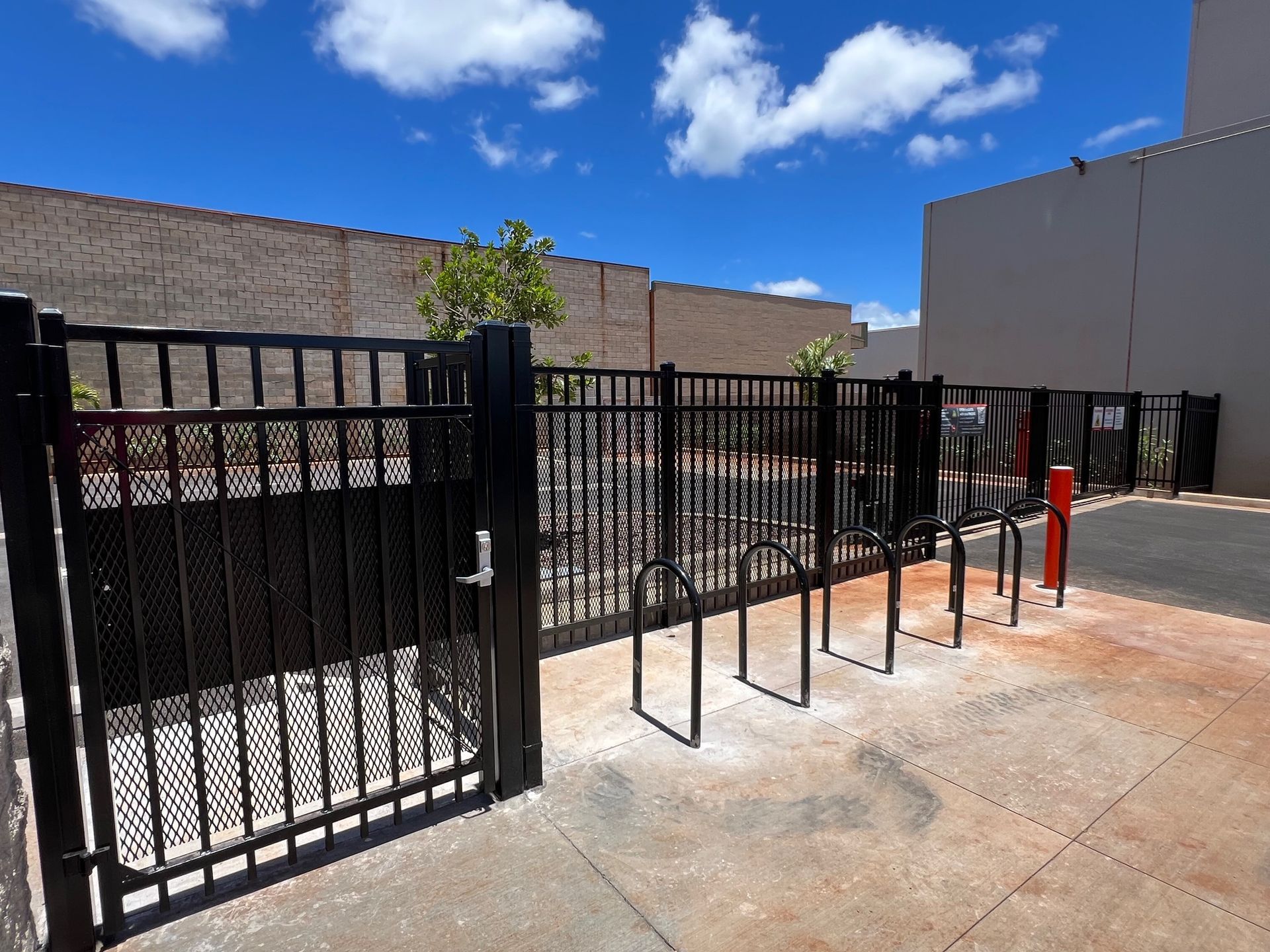 Black metal fence encloses a bike rack area. Blue sky with clouds. Brick wall in the background.