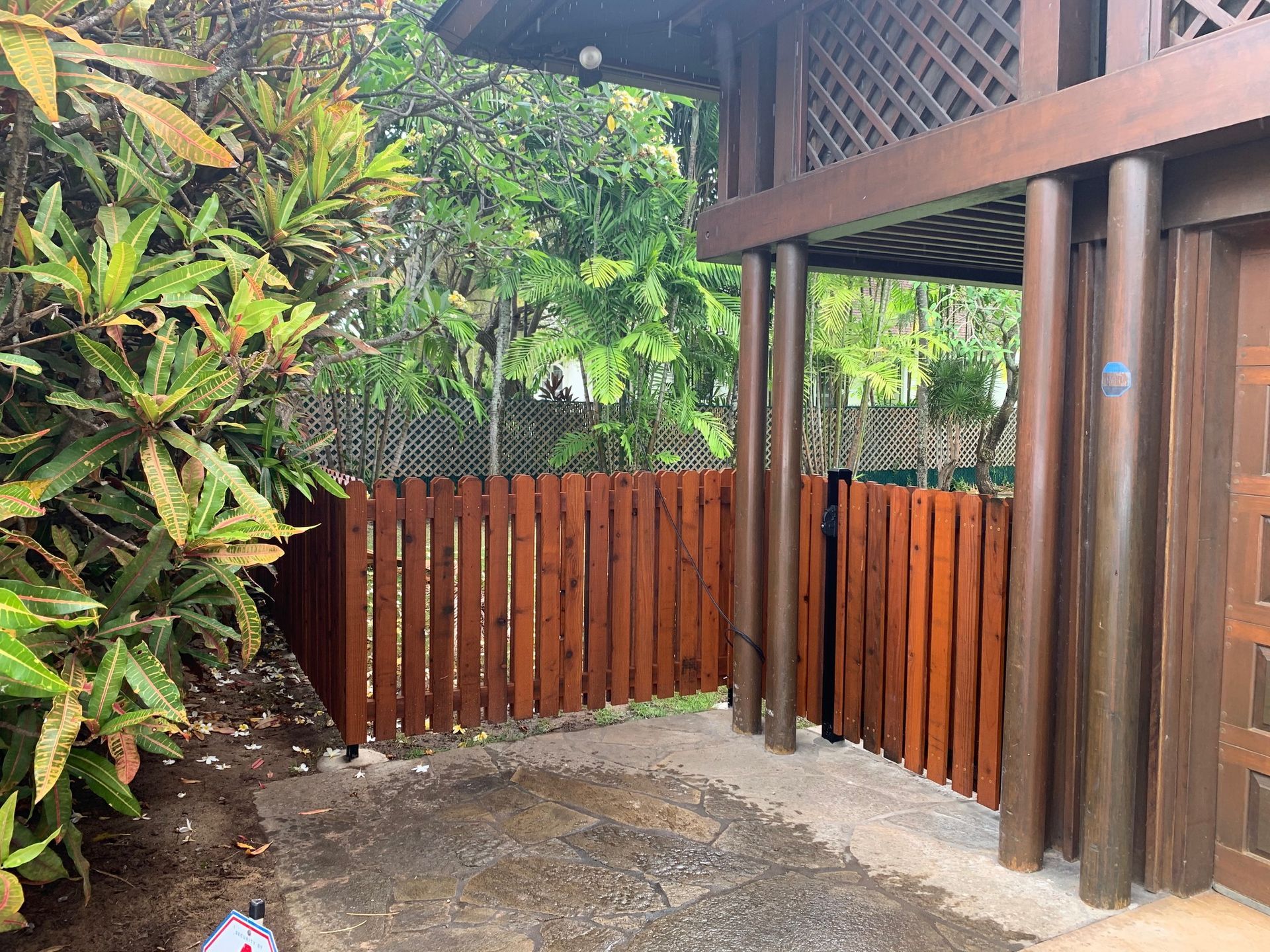 Brown wooden fence and gate next to a building and greenery.