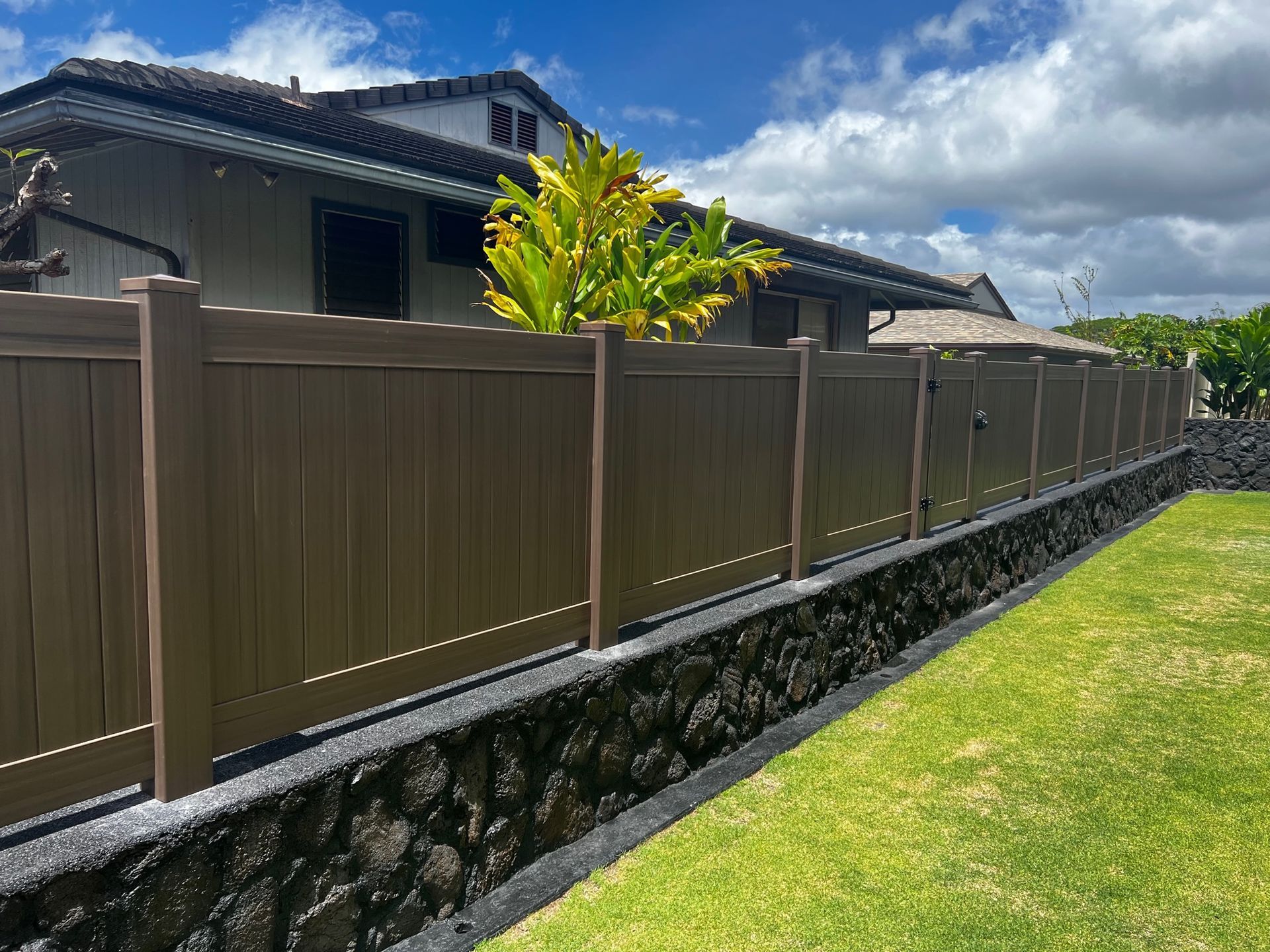 Brown fence atop a stone wall, bordering a grassy lawn in front of a house, under a cloudy blue sky.