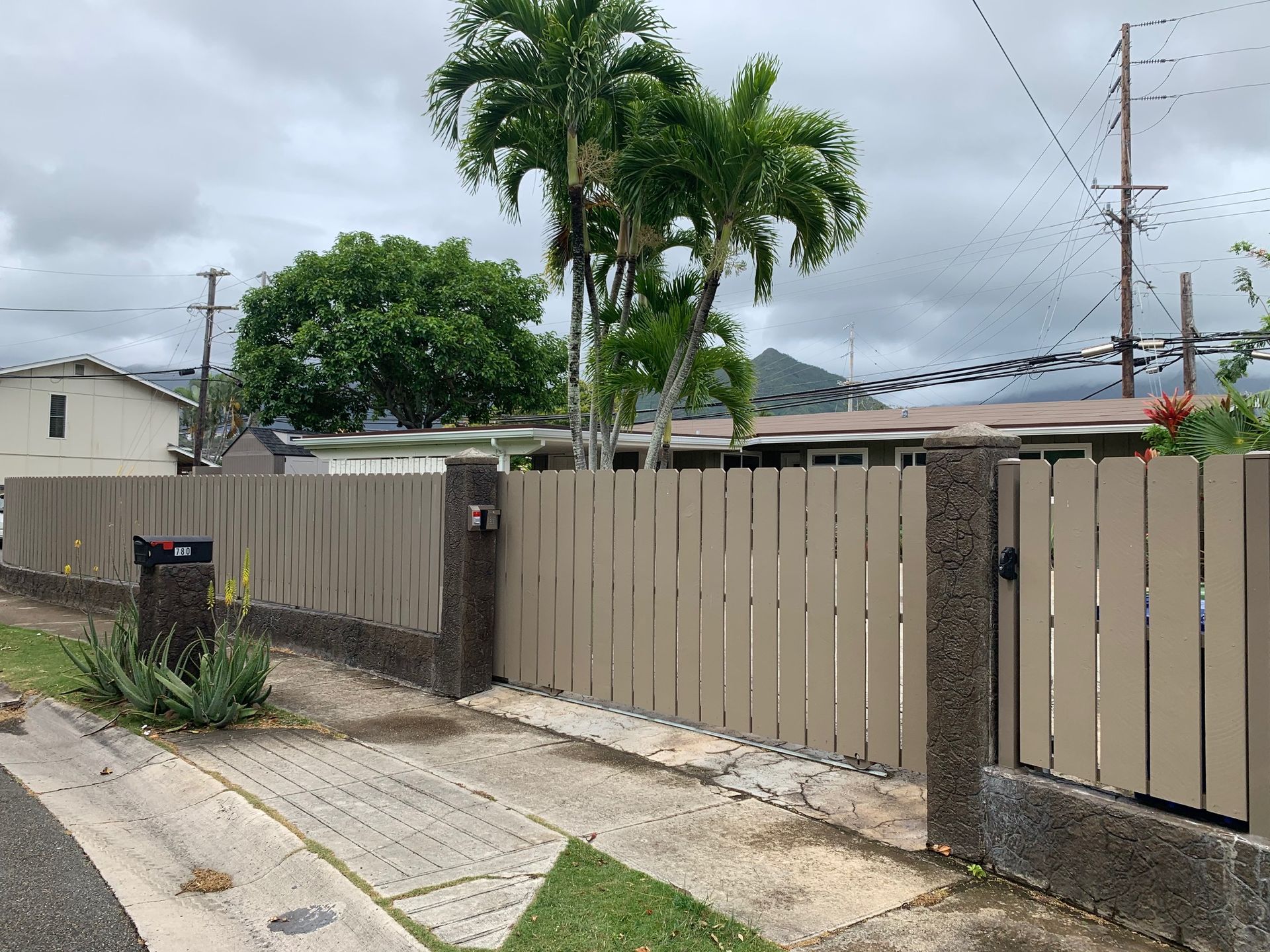 Tan wooden fence surrounds a house with palm trees, set against a cloudy sky.